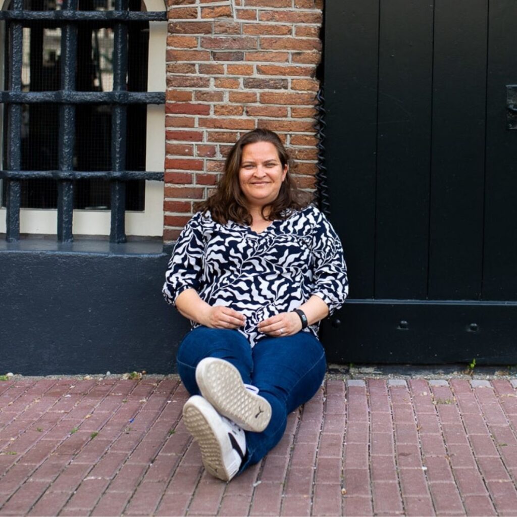 Elvira zit op de grond voor een grote deur, kijkt recht de camera in.